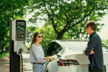 Doğal orman ya da milli parktaki araba yolculuğu sırasında elektrikli araba için şarj olan güneş gözlüğü takan sevimli genç bir çift. Tatil ve tatil boyunca çevre dostu seyahat. Yükselt