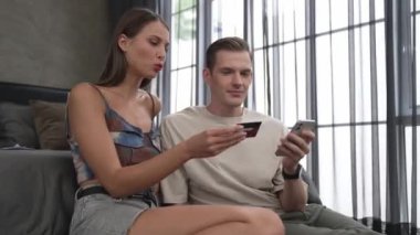 Genç çift, kredi kartıyla ödemek için internet üzerinden ödeme uygulaması ve dijital cüzdan kullanarak yatak odasında oturuyor. Mobil internet üzerinden alışveriş ve modern alımlar. Düzenle