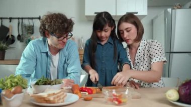 Çekici beyaz anne ve baba Asyalı kızıyla taze yemek pişirmek için birlikte vakit geçiriyorlar. Enerjik anne ve baba mutfakta salata yapmak için taze sebze hazırlıyor. Sağlıklı yiyecekler. Pedagoji.