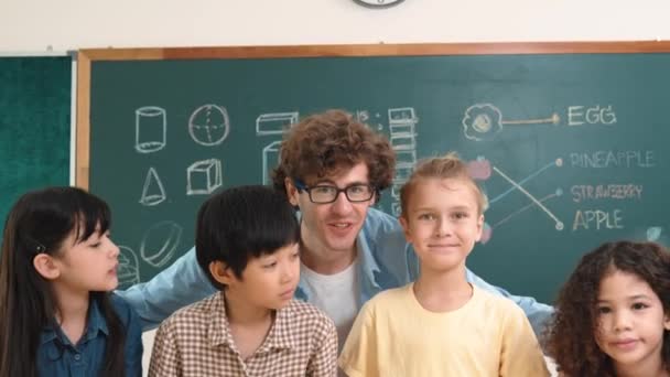 教室でカメラを見ている白人スマートな先生と多様な学生 ハッピーな子供たちのグループは笑顔で インストラクターはブラックボードの前で励まし 立っています 教育について ペダゴロジー — ストック動画
