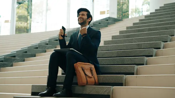 Successful business man celebrate successful project while sitting at stairs. Smart project manager getting new gob, getting promotion, increasing sales while calling friends by using phone. Exultant.