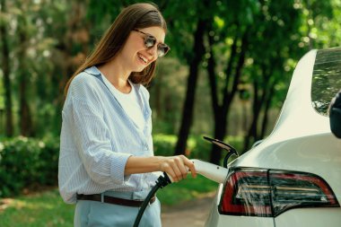 Yolculuk sırasında doğal orman ya da milli parktaki elektrikli araba için şarj olan genç bir kadın. Tatil ve tatil boyunca çevre dostu seyahat. Yükselt
