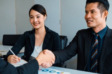 İş adamları konsepti kabul ediyor. Asyalı iş adamı ofis toplantı odasında başka bir iş adamıyla tokalaşıyor. Genç Asyalı sekreter onun yanında oturuyor. uds