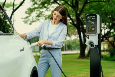 Açık yeşil şehir parkı manzaralı EV şarj istasyonundan genç kadın EV elektrikli araçları şarj ediyor. Çevre dostu şehir ulaşımı ve çevre dostu EV araba seyahati. Yükselt