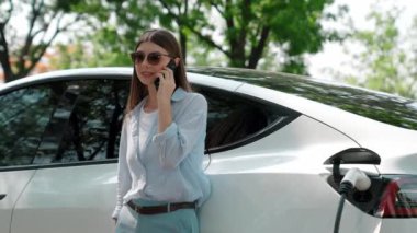 Doğal orman ya da milli parktaki araba yolculuğu sırasında elektrikli araba pili şarj ederken kadın telefonda konuşuyor. Tatil ve tatil boyunca çevre dostu seyahat. Yükselt