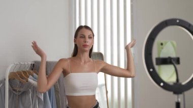 Genç sosyal medyum kadın moda videosu yapıyor. Blogcu kameraya selfie çubuğu ve ışık halkası kullanarak gülümserken, aynı zamanda ikna edici çevrimiçi kıyafetlerin izleyicilere veya takipçilere vlog satmasını sağlıyor. Düzenle