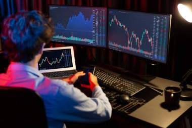 Akıllı telefonu elinde tutan genç yatırımcı hisse senedi tüccarı bilgisayardaki dinamik döviz kuru verileriyle karşılaştırıyor ve güncel pazar grafiğinde Neon Light Modern Ofis 'te gerçek zamanlı olarak. Toplayıcı.