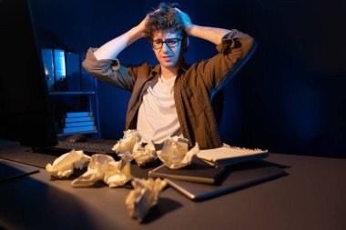 Aşırı çalışan projeyle uğraşan stresli yaratıcı yönetici buruşmuş kağıtların etrafını sararken gece vakti modern ofisteki gündelik gömlekle dizüstü bilgisayar ekranına e-posta göndermeyi bekliyor. Toplayıcı.