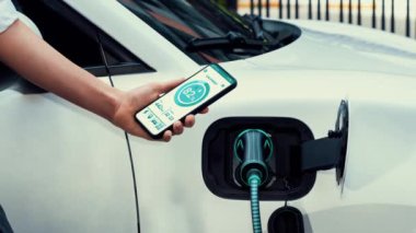 Akıllı telefon akü durumu arayüzünü elinde tutan iş kadını akıllı EV mobil uygulama ile otomobil parkındaki şarj istasyonundan elektrik şarj eden çevre dostu EV araba. İnceleyin