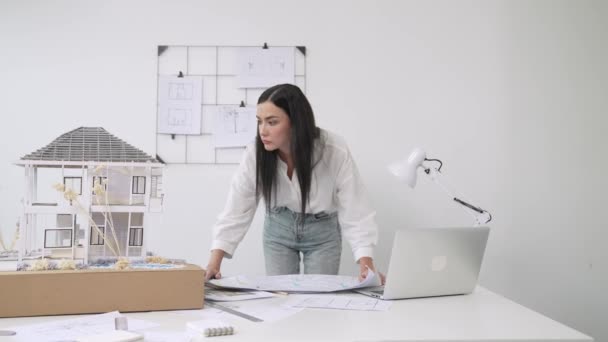 年轻美丽的高加索建筑师把蓝图摆到桌上 并与房屋模型进行比较 同时在现代办公室仔细检查带有条纹的蓝图的模型建设 纯洁无瑕 — 图库视频影像