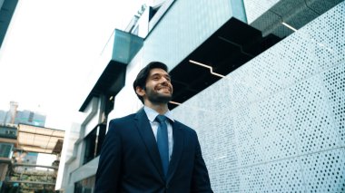 Takım elbiseli ve gülümseyen zeki bir iş adamı beyaz modern mimari binasının yanında sokakta yürürken. Şehir merkezinde çekici, yakışıklı bir yönetici gökdelen gibi görünüyor. Uzayı kopyala Sevinçli..