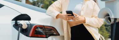 Genç bir kadın EV otomobil şarj istasyonu Green City Park 'ta elektrik ödemek için akıllı telefonu kullanıyor. Modern çevre ve EV araçlı sürdürülebilir şehir yaşam tarzı. Panorama Çıkarı