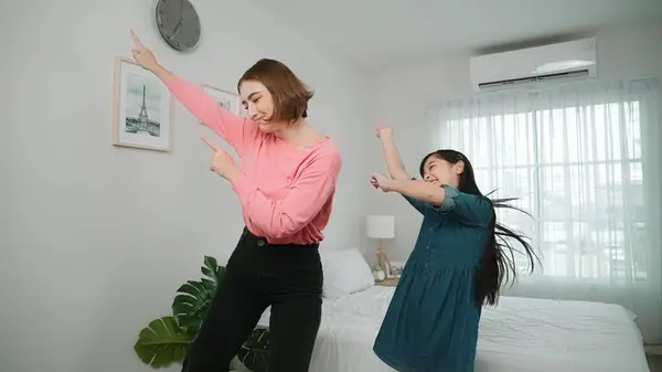 Happy attractive mother dancing to music together with asian daughter at bedroom. Caucasian mom dance or exercise while cute child move to music while jump to bed. Family recreation concept. Pedagogy.