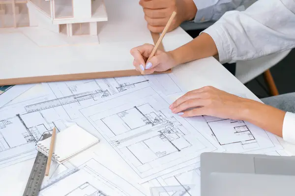 Top view image of professional beautiful young architect hand drafts blueprint while asian cooperative coworker measure house model on table with house model and blueprint scatter around. Immaculate.