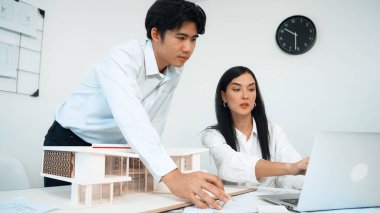 Profesyonel Asyalı erkek mimar ev modelinin uzunluğunu ölçmek için cetvel kullanırken, beyaz tenli genç meslektaşı ev modeliyle toplantı masasındaki verileri analiz etmek için dizüstü bilgisayar kullanıyor. Tertemiz..
