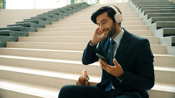 Smiling business man listening music while looking at mobile phone. Project manager holding phone while enjoy hip-hop song at stairs. Caucasian leader listen relax sound while plan project. Exultant.