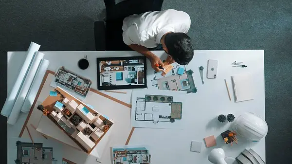 Top view of business man using tablet plan house design while sitting at meeting table with blueprint and equipment scatter around. Aerial view of civil engineer working at blueprint. Alimentation.