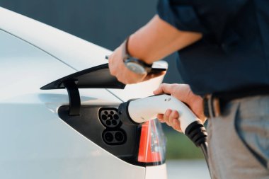 Genç adam, şehrin ticari otoparkındaki şarj istasyonundan elektrikli araba aküsünü şarj etmek için EV şarj aleti koymuş. Sürdürülebilir çevre dostu şehir yolculuğu için şarj edilebilir EV araba. Yararlı