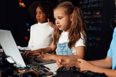Beyaz önlüklü akıllı kız BTMM sınıfında dizüstü bilgisayar kullanarak robotik teknolojiyi öğreniyor. Mavi tişörtlü öğrenci anakart öğretirken zeki öğrenciler kod okumaya çalışıyor. Etkinlik.