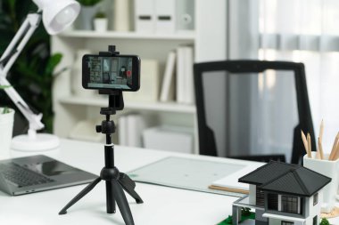 Akıllı telefonun beyaz masanın üzerinde durduğu mimari kayıt için tüm ekipman setleri modern ofiste canlı yayın yapan dizüstü bilgisayar, kağıt, model ev ve kalemden oluşur. Toplayıcı.