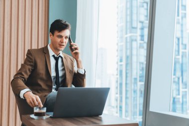 Gökdelen manzaralı pencerenin yanında otururken yakışıklı iş adamının müdürle telefon görüşmesi yaptığı yakın plan. Yönetici, telefon ve dizüstü bilgisayarla konuşuyor. Önüne bak. Süslü.