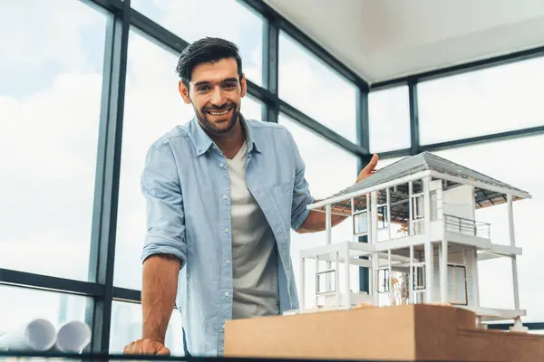 Portrait of smart engineer in casual outfit smiling at camera while inspect house model. Skilled architect looking at camera and standing near house model, project plan, architectural model. Tracery.