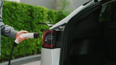 Genç adam yeşil sürdürülebilir şehir açık hava yaz bahçesinde EV elektrikli otomobilini yeniden şarj ediyor kentsel sürdürülebilirlikte gençlerin yaşam tarzı yeşil temiz şarj edilebilir enerji elektrikli araç iç organları
