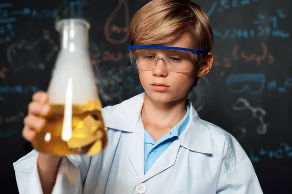 Blonde hair schoolboy in laboratory wear lab coat stand and learn science of chemistry technology in STEM class. The student pour liquid down to glass beaker. On the table put many flask. Erudition.
