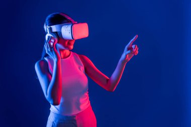 Siberpunk neon ışıklı cihazlarla çevrili zeki bir kadın metaevrene bağlı VR kulaklık takıyor, gelecekçi siber uzay teknolojisi. Sanal objeyi işaret eden bir kadın. Halüsinasyon.