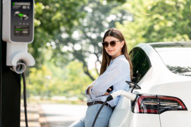 Yolculuk sırasında doğal orman ya da milli parktaki elektrikli araba için şarj olan genç bir kadın. Tatil ve tatil boyunca çevre dostu seyahat. Yükselt