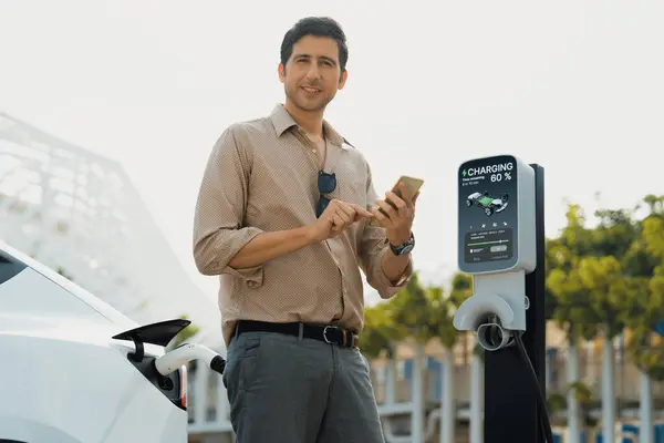 年轻人在公共电动车收费站绿城公园用智能手机支付电费 现代环境和可持续城市生活方式与电动汽车 — 图库照片