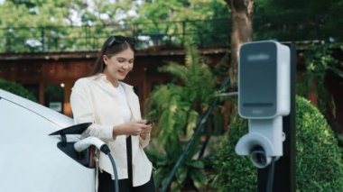 İlkbahar bahçesindeki açık kafede çalışan EV elektrikli araba şarjı, yeşil şehir sürdürülebilirliği ve çevre dostu EV otomobili için genç bir kadınla sürdürülebilir şehir seyahati elektrik ödüyor. Yararlı