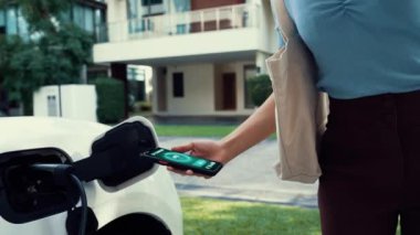 Modern yaşam tarzı bir kadın EV şarj cihazını ev şarj istasyonuna yerleştirir ve akıllı telefondan EV batarya durumunu etkinleştirir. Gelecekçi enerji sürdürülebilirliği ve elektrikli araba. İnceleyin