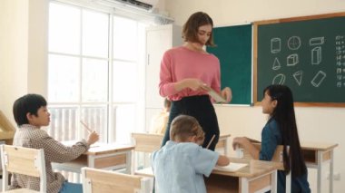 Zeki Asyalı öğrenci, ilkokulda öğretmen yürürken ve ödevlerini kontrol ederken matematik sorusunu sorarak elini kaldırdı. Öğretmen not defterini alırken soruyu cevaplıyor. Pedagoji.