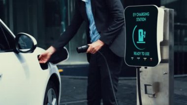 İşadamı EV şarj aletini şehir merkezindeki EV şarj istasyonundan ya da otoparktan şarj etmek için takıyor. Çevre dostu şarj edilebilir araba şehir yaşamında alternatif enerji kullanıyor. İnceleyin.