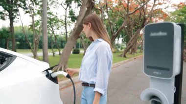 Yavaş çekimde genç bir kadın sonbaharda ulusal parktaki EV şarj istasyonunda EV elektrikli arabaları şarj ediyor. Tatil ve sonbahar sezonunda çevre dostu arabalarla seyahat. Yararlı