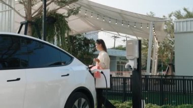 Genç kadın otoparktaki şarj istasyonundan EV araba aküsünü alışveriş çantasıyla şarj ediyor. Modern kadın, şehir içi seyahat tarzında çevre dostu elektrikli araçla alışverişe gider. Yararlı