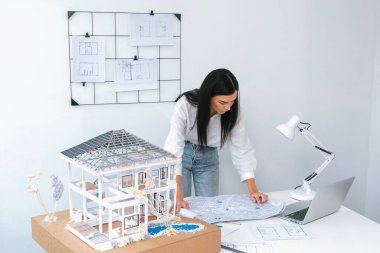 Genç, beyaz, güzel bir mimarın portresi toplantı masasına yerleştirilen ev modeli, plan ve dizüstü bilgisayarın yapısını karşılaştırmaya odaklanıyor. Şah, teftiş, tahmin konsepti. Tertemiz..