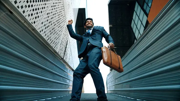 Low angle view of young smiling business man dance between building at center point. Skilled executive manager wearing suit and suitcase and moving to music. Happy investor moving to music. Exultant.