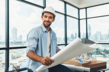 Ev modelinde proje planını ve gökdelenli mimar ekipmanlarını okurken kameraya bakan profesyonel mimar mühendisinin portresi. Mimarlık stüdyosuyla çevrili. İzleme.