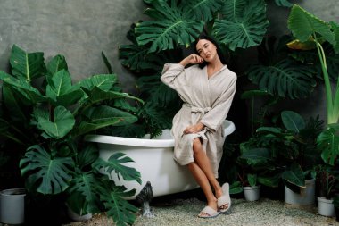 Modern otelde küveti olan tropikal ve egzotik bir spa bahçesi ya da yemyeşil yeşillik arka planıyla çevrili rahat ve sağlıklı yaşam tarzının tadını çıkaran bornozlu bir kadınla tatil köyü. Blithe