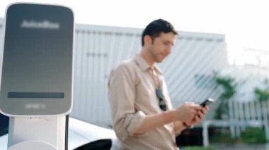 Ağır çekimde çalışan adam, alışveriş merkezinin otoparkındaki kamu EV şarj istasyonunda elektrik için akıllı telefon kullanıyor. EV aracıyla modern çevre ve sürdürülebilir kentsel yaşam tarzı