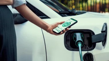 Akıllı telefon akü durumu arayüzünü elinde tutan iş kadını akıllı EV mobil uygulama ile otomobil parkındaki şarj istasyonundan elektrik şarj eden çevre dostu EV araba. İnceleyin