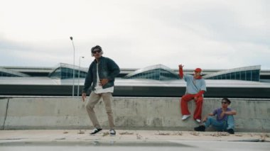 Yakışıklı break dansçı, Asyalı arkadaşının tezahürat yaptığı serbest stil hareketinden hoşlanır. Hippi kulaklıktan müzik dinler ve kameraya bakar. Sokak dansı. Açık hava sporu 2024. Yumuşak bir şekilde..