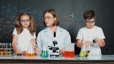 Öğretmen tavsiye verirken beyaz çocuk kimyasal sıvıyı karıştırıyor. Profesyonel eğitmen labaratuar kıyafeti giymiş, renkli solüsyonla dolu deney kabıyla sofrada çeşitli öğrencileri arıyor. Etkinlik.