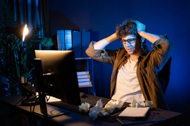 Aşırı çalışan projeyle uğraşan stresli yaratıcı yönetici buruşmuş kağıtların etrafını sararken gece vakti modern ofisteki gündelik gömlekle dizüstü bilgisayar ekranına e-posta göndermeyi bekliyor. Toplayıcı.