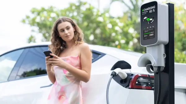 集中充电站充电电动车的模糊背景下的现代妇女使用智能手机 Ev技术用于跟踪能源使用情况以优化电池充电 — 图库照片