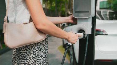Genç bir kadın yazın yeşil sürdürülebilir şehir bahçesinde EV elektrikli arabasıyla seyahat ediyor. Kentsel sürdürülebilirlik yaşam tarzı yeşil temiz yeniden şarj edilebilir enerji elektrikli BEV araç iç organları