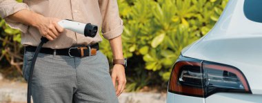 Genç adam yeşil şehir parkındaki şarj istasyonundan elektrikli araba aküsünü şarj ediyor. Sürdürülebilir çevre dostu şehir yolculuğu için şarj edilebilir EV araba. Panorama Çıkarı