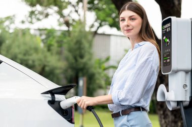 Açık yeşil şehir parkı manzaralı EV şarj istasyonundan genç kadın EV elektrikli araçları şarj ediyor. Çevre dostu şehir ulaşımı ve çevre dostu EV araba seyahati. Yükselt